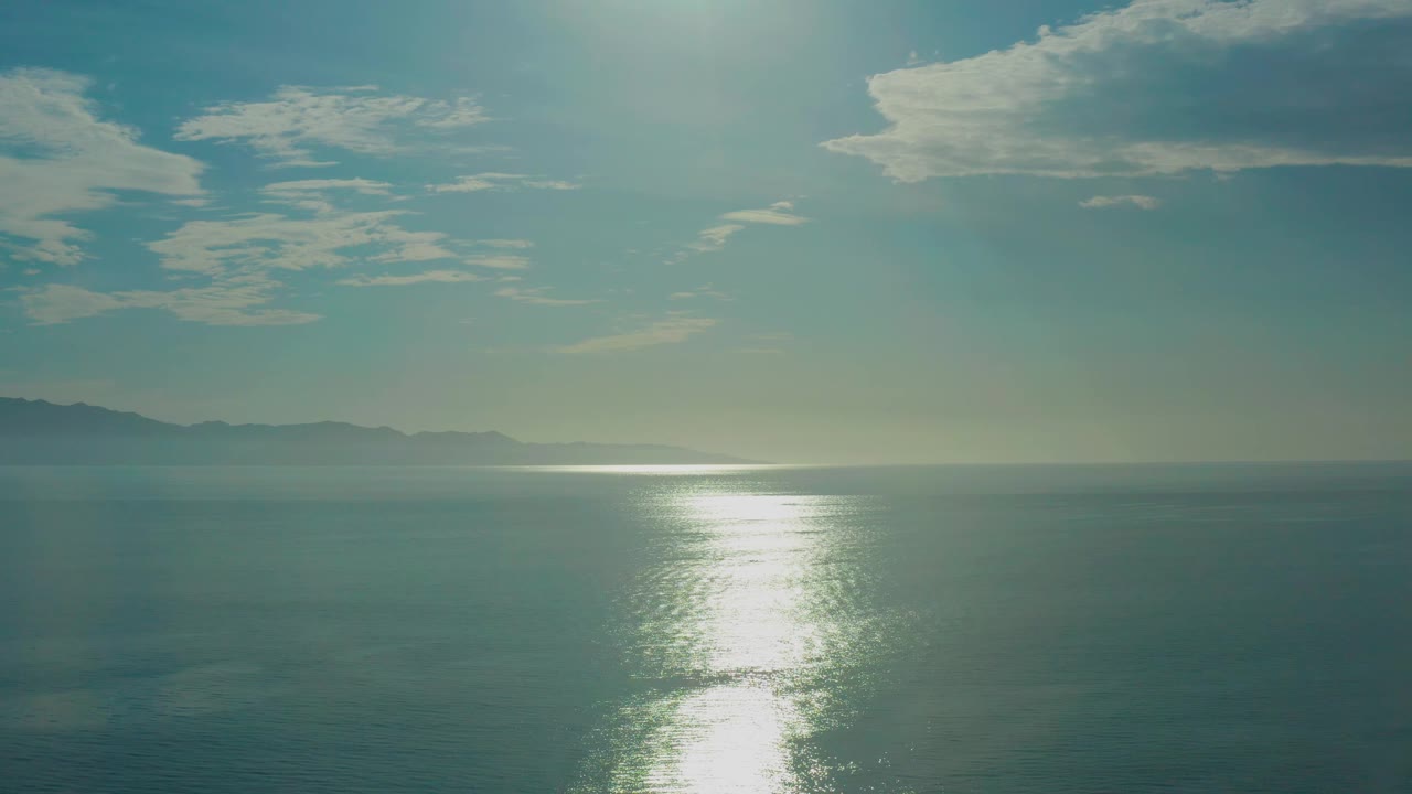 At 0:02, the reflected sun patch brightens the midwater while the hazy left coastline remains low on the horizon near El Sargento La Ventana, Mexico, with no visible people.