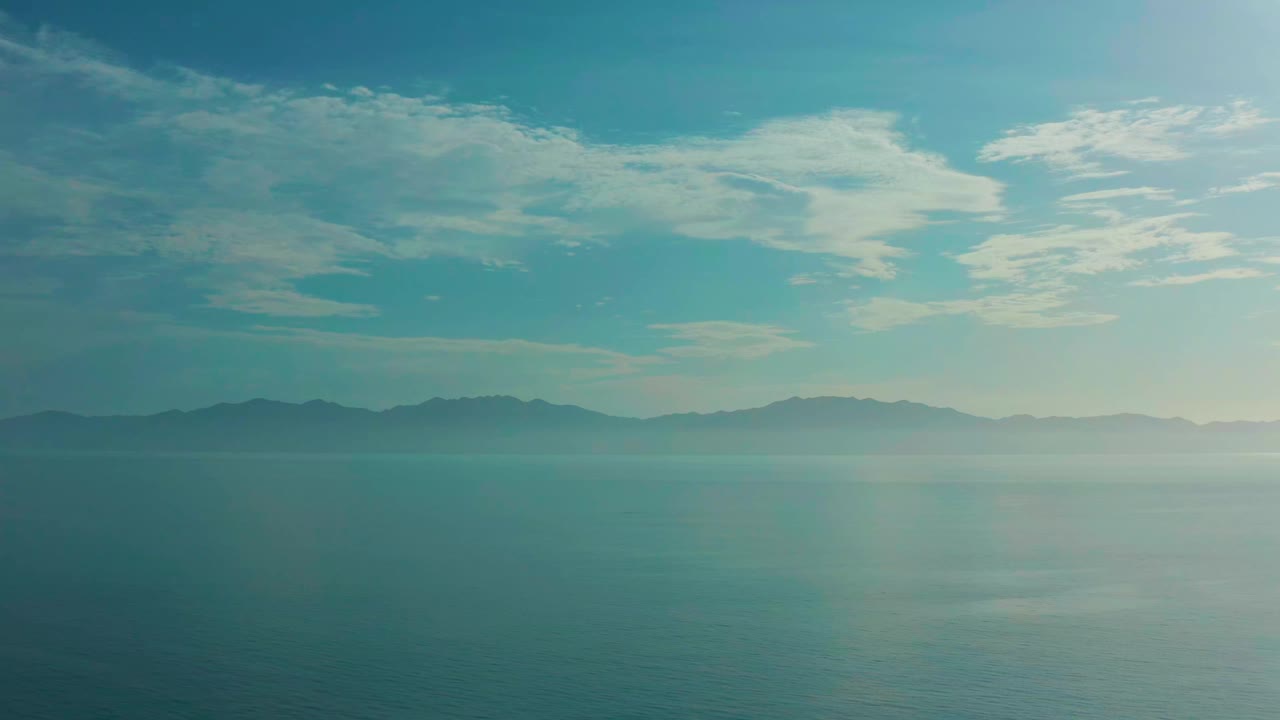 A wide frame shows blue-green water in the foreground, pale mist near the horizon, and layered mountains beneath bright clouds in El Sargento La Ventana, Mexico, with no visible people.