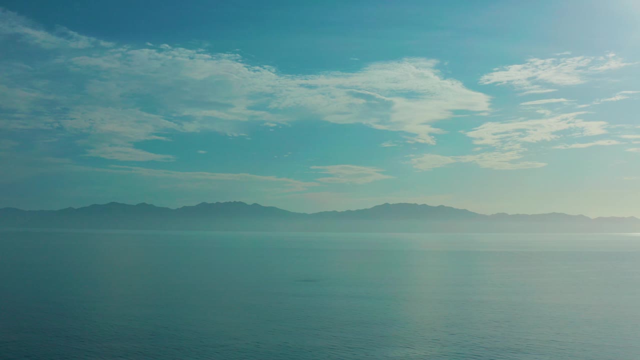 At 0:03, broad water fills the lower frame in El Sargento La Ventana, Mexico, with a low mountain band across the distance, wispy clouds above, and no visible people or vessels.