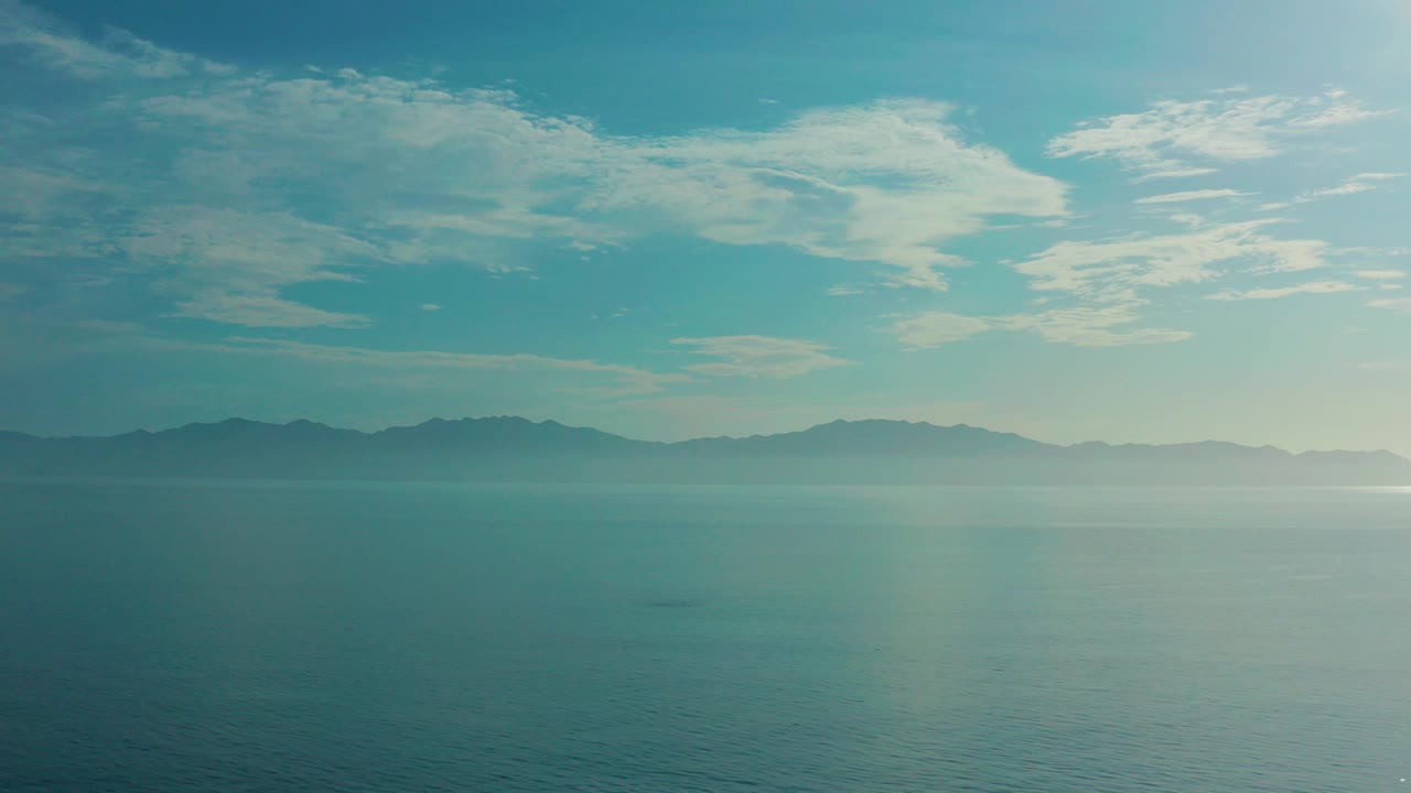 Opening frame over calm blue water near El Sargento La Ventana, Mexico, with hazy land on the horizon, thin cloud cover, and no visible people.