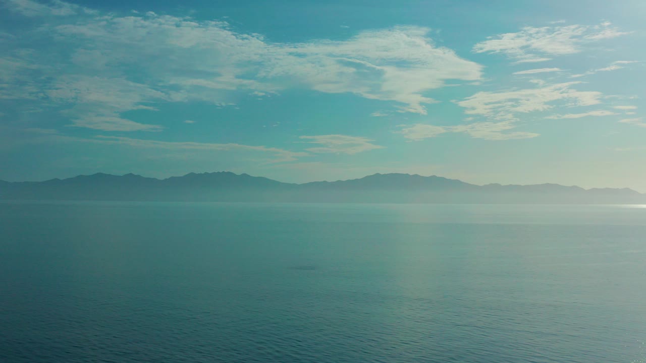 Wide blue-green ocean view in El Sargento La Ventana, Mexico, with a dark mountain line on the horizon, pale clouds overhead, and no visible people.