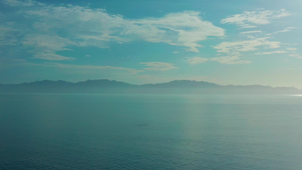 High wide ocean frame from El Sargento La Ventana, Mexico, with layered mountains stretching across the background, scattered white clouds, and no visible people.