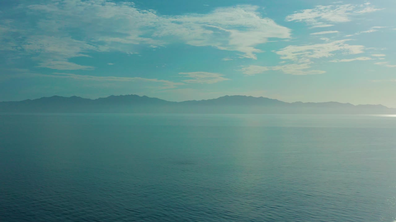 Opening frame over teal water in El Sargento La Ventana, Mexico, with distant hazy land on the horizon, thin cloud cover above, and no visible people.