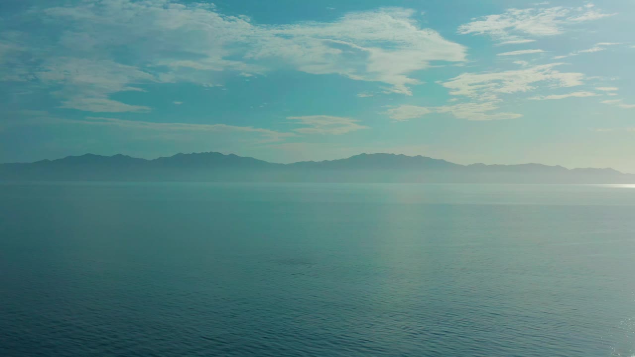 A wide view across calm teal water toward a muted mountain shoreline in El Sargento La Ventana, Mexico, with pale cloud bands and no visible people.