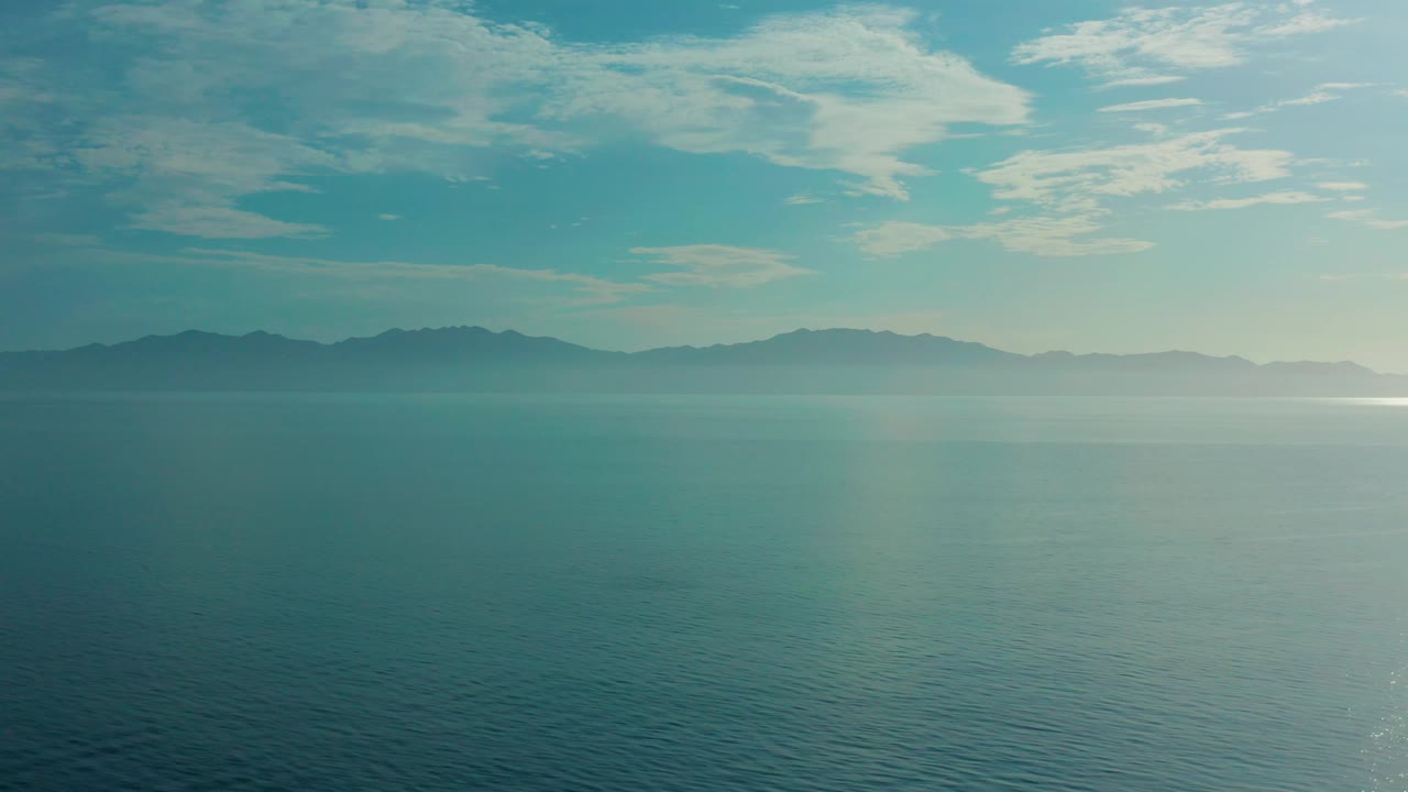 Mid-clip frame looking across blue-green water toward a misted strip of land in El Sargento La Ventana, Mexico, with layered clouds above and no visible people.
