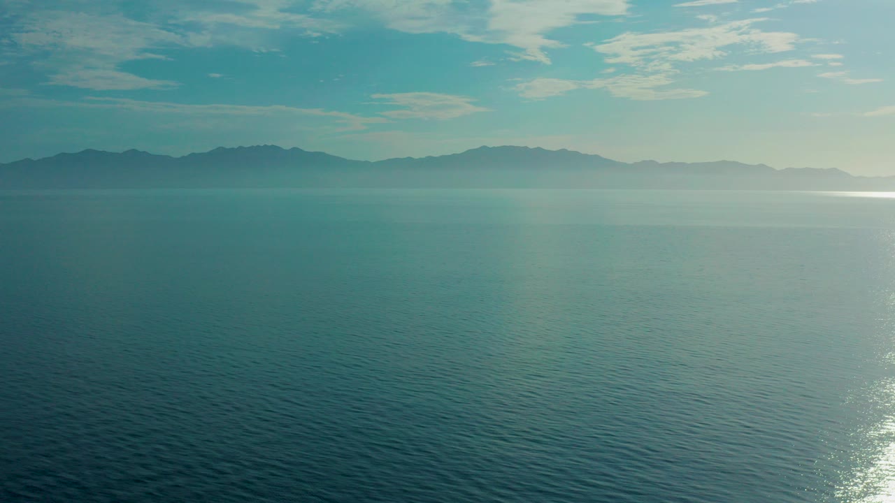 Wide aerial view over blue ocean near El Sargento La Ventana, Mexico, with hazy mountains on the horizon, thin clouds above, and no visible people.