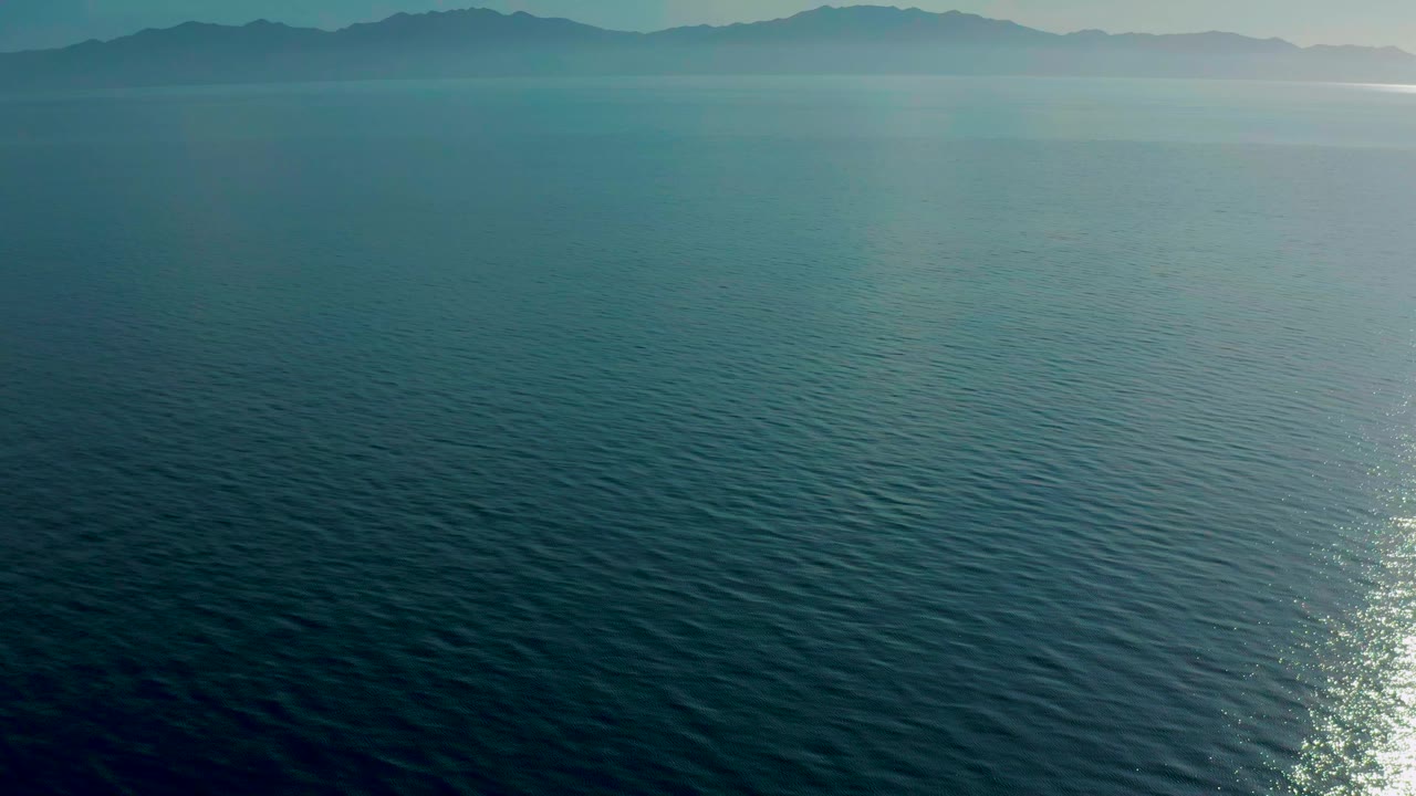 Aerial view over teal-blue sea toward a hazy mountain horizon in El Sargento La Ventana, Mexico, with sun reflection low in frame and no visible people.