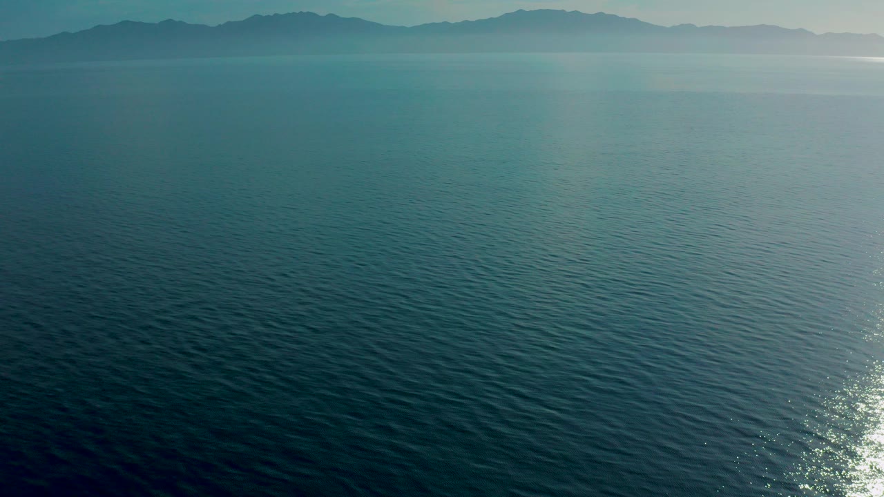 Aerial view over blue-green ocean at El Sargento La Ventana, Mexico, with hazy mountains along the horizon and no visible people.