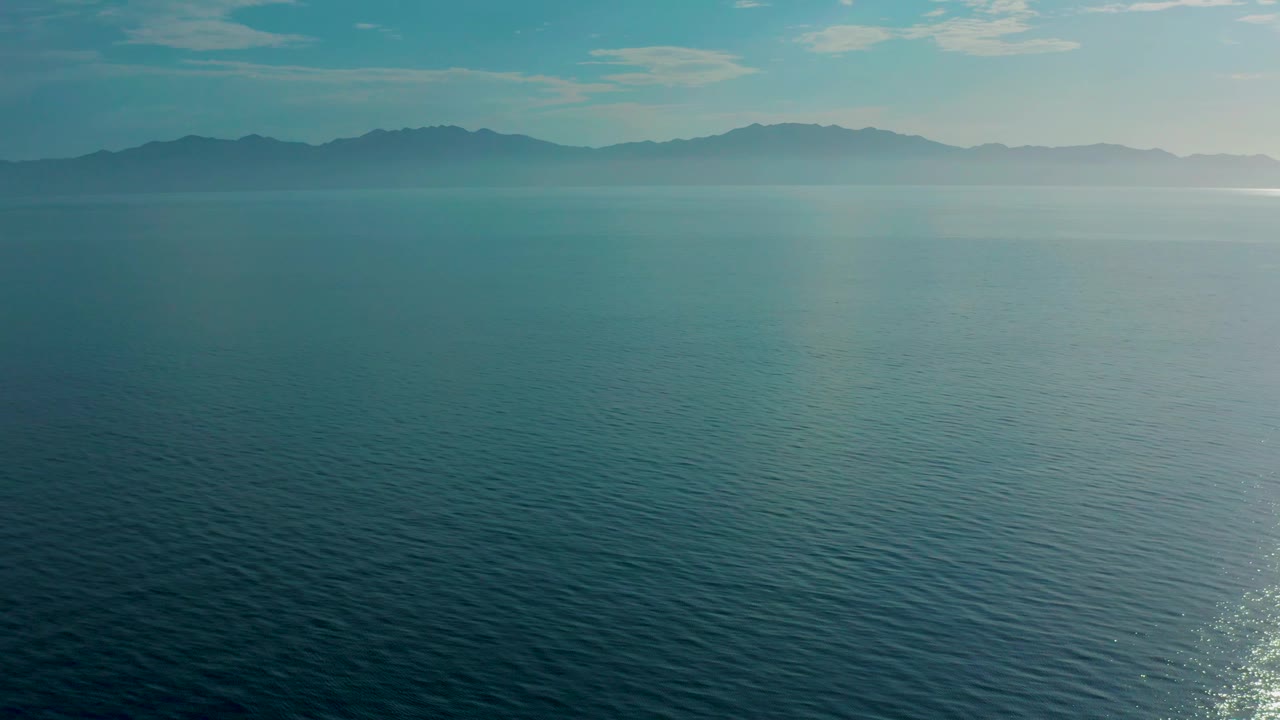 Wide aerial view of teal open water with mountain silhouettes across the horizon and a bright patch of sun glare near the right edge, with no visible people.