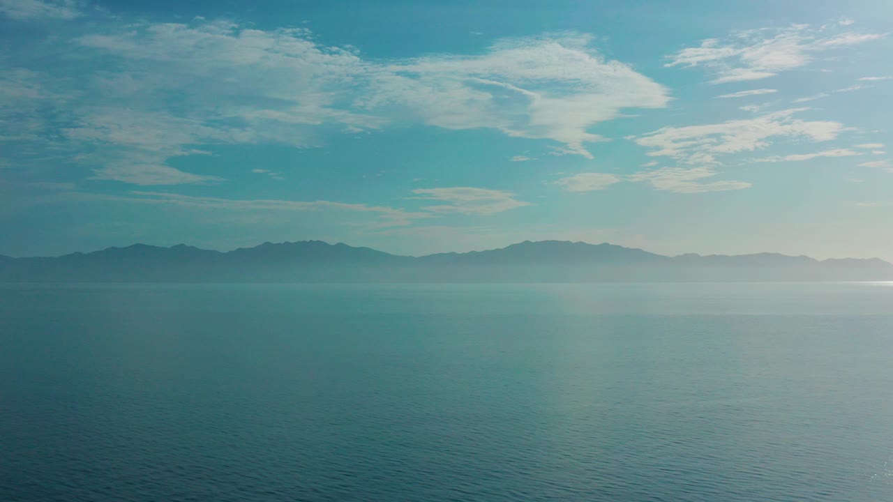 Opening view of blue ocean and a dark mountain horizon beneath high pale clouds in El Sargento La Ventana, Mexico, with no visible people.
