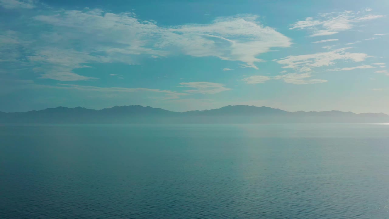 A steady wide aerial view shows calm blue-green water, silhouetted mountains, and a band of bright clouds over El Sargento La Ventana, Mexico, with no visible people.