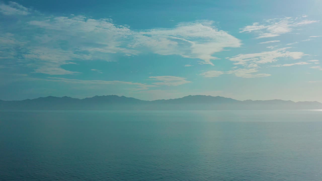 Mid-clip frame with empty blue water below, dark mountains across the horizon, and streaked white cloud cover over El Sargento La Ventana, Mexico, with no visible people.
