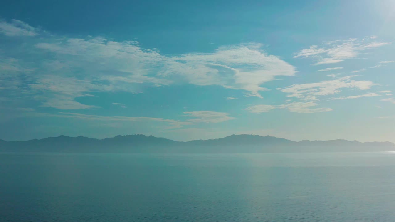 Calm sea and distant mountain ridge under thin white clouds.