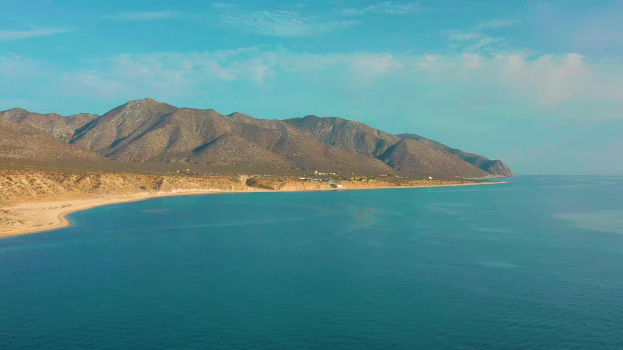 Wide aerial view of calm blue water meeting the pale coast of El Sargento La Ventana, Mexico, with desert-toned mountains behind it and no visible people.