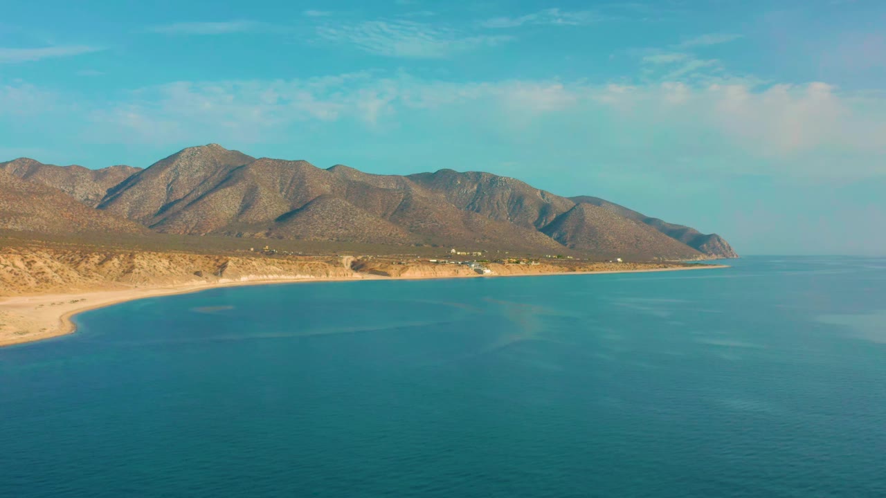 Second aerial frame in El Sargento La Ventana, Mexico, showing calm blue water below the sandy shoreline, low scrub terrain, a long brown mountain ridge, and no visible people.