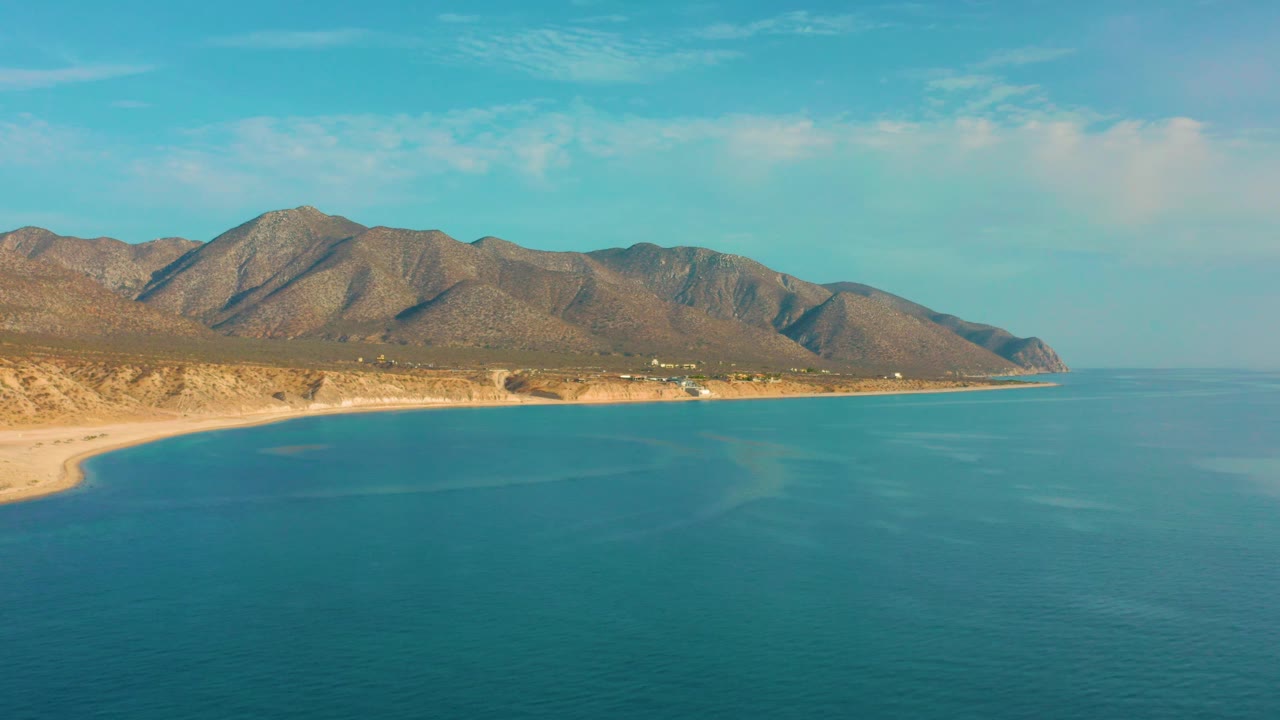 Wide aerial coastline with turquoise water, sandy beach, and rugged hills.