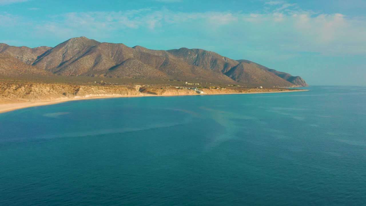Opening aerial frame over El Sargento La Ventana, Mexico, with blue sea in the foreground, a sandy coast below brown mountains, scattered clouds, and no visible people.