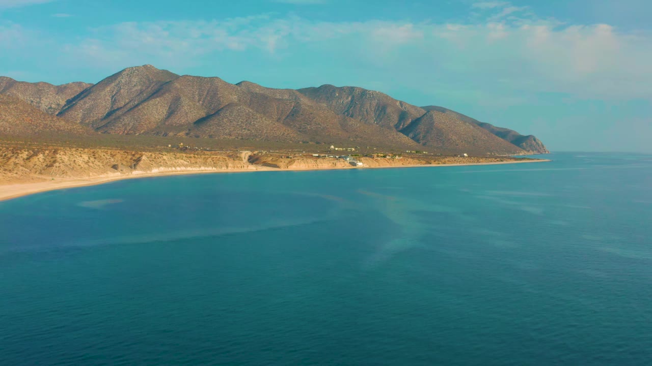 Early aerial frame from El Sargento La Ventana, Mexico, showing turquoise water across the foreground, a narrow sandy beach, sunlit mountain slopes, and no visible people.