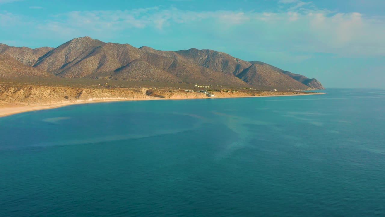 Aerial view in El Sargento La Ventana, Mexico, with pale underwater color patches in the blue sea, a slim beach, rugged brown mountain ridge, and no visible people.