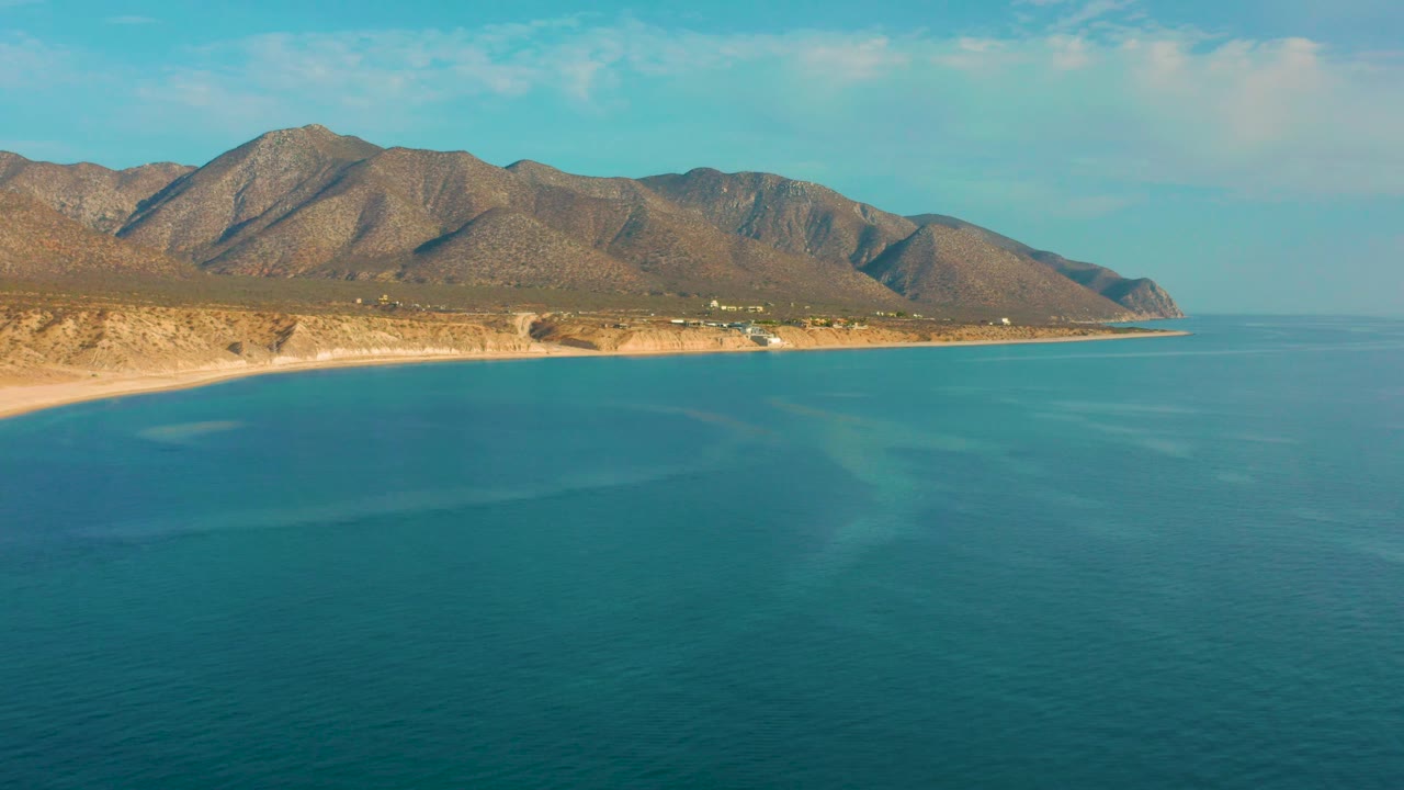 Mid-clip frame over El Sargento La Ventana, Mexico, with broad blue water in front, a curving sandy shore, arid mountain slopes, soft cloud cover, and no visible people.