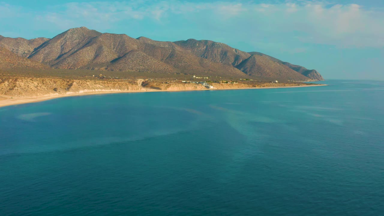 Early frame showing teal water in the foreground, a pale beach along the left edge, distant mountain slopes, and no visible people in El Sargento La Ventana, Mexico.