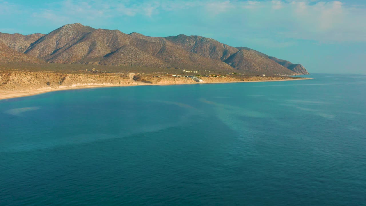 Mid-clip frame with the sandy beach more prominent at left, blue water across the foreground, dry mountains behind, and no visible people in El Sargento La Ventana, Mexico.