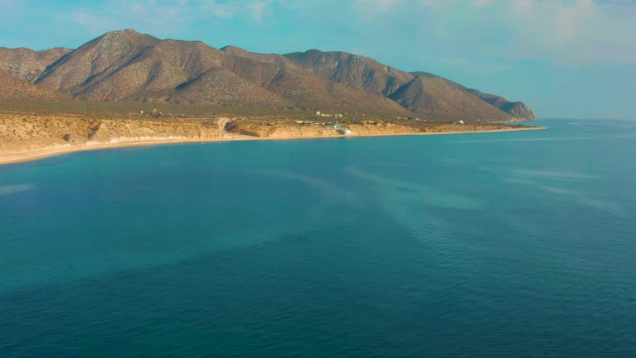 A wide aerial view in El Sargento La Ventana, Mexico, shows turquoise water reaching a narrow sandy coast below brown mountains, with no visible people.