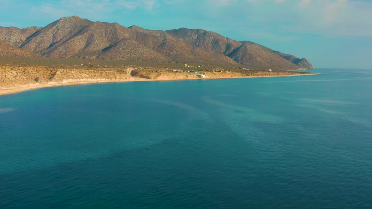 Bright blue sea fills most of the frame in El Sargento La Ventana, Mexico, with a pale shoreline and mountain ridge along the upper third, and no visible people.