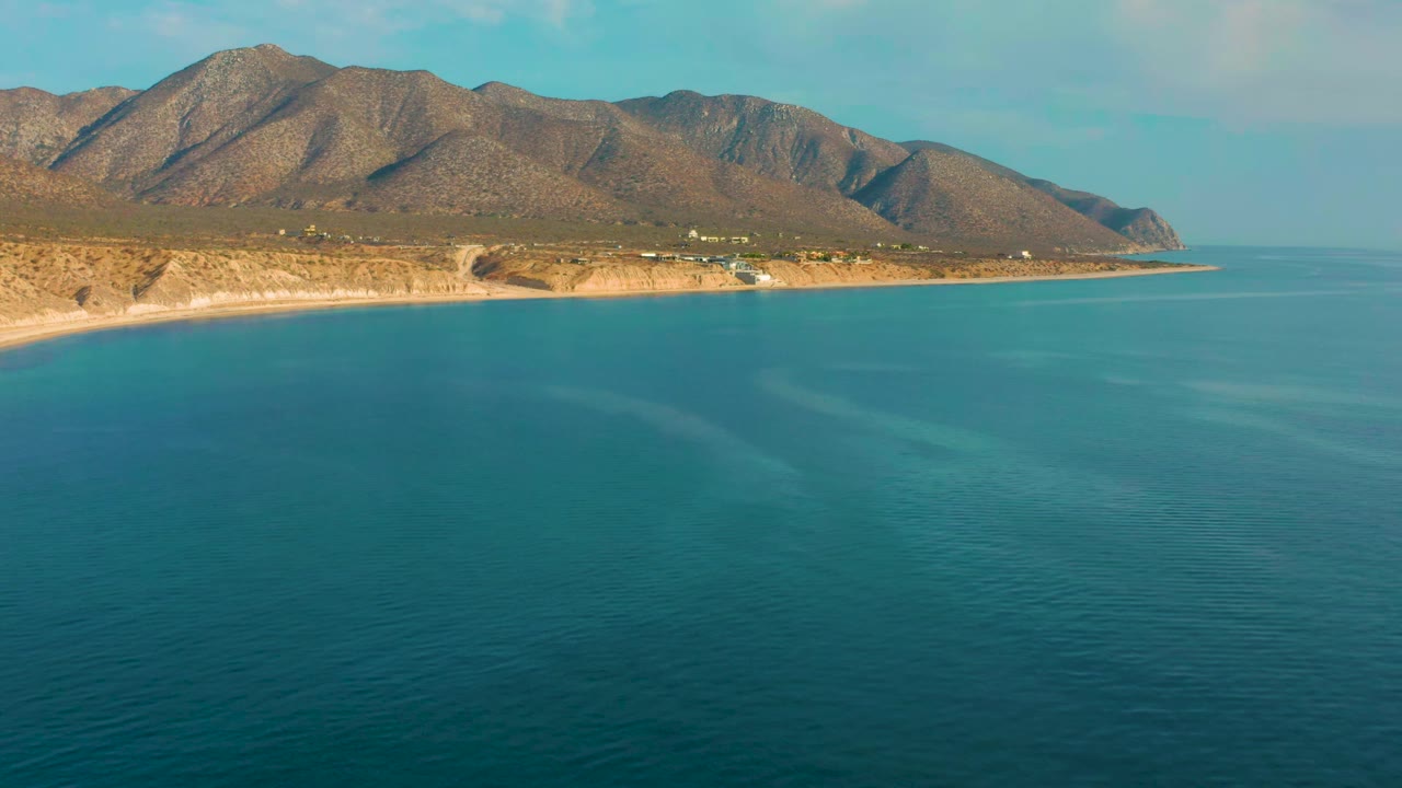 Second frame of the El Sargento La Ventana coastline, showing turquoise water across the foreground, a narrow pale beach, scattered small buildings, mountains, and no visible people.