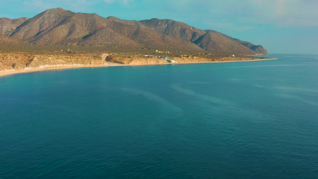 Mid-clip frame over El Sargento La Ventana, Mexico, with bright blue ocean filling the lower frame, sunlit tan shore, brown mountains, and no visible people.