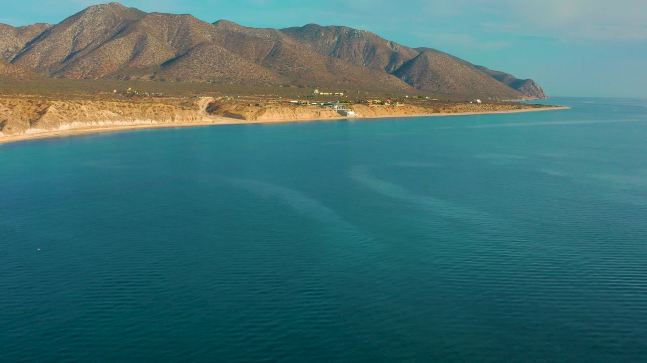 Opening aerial frame over El Sargento La Ventana, Mexico, with blue ocean in front, a pale shoreline below dry mountains, and no visible people.