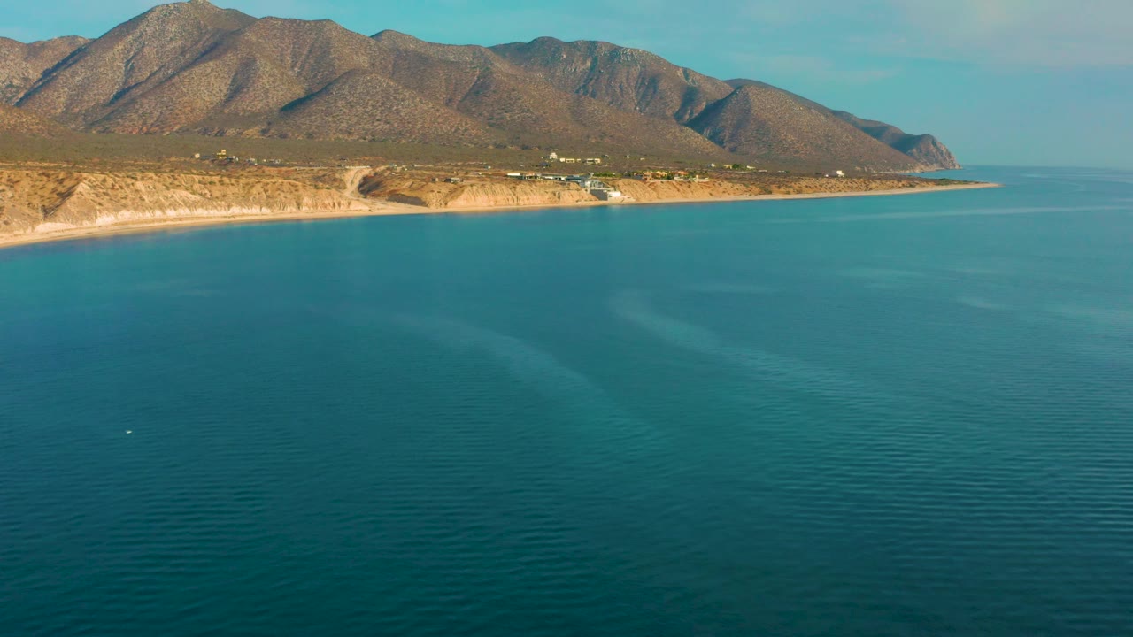 High coastal frame from El Sargento La Ventana, Mexico, with the mountain range stretching across the top, a narrow shore band in the middle, blue sea below, and no visible people.