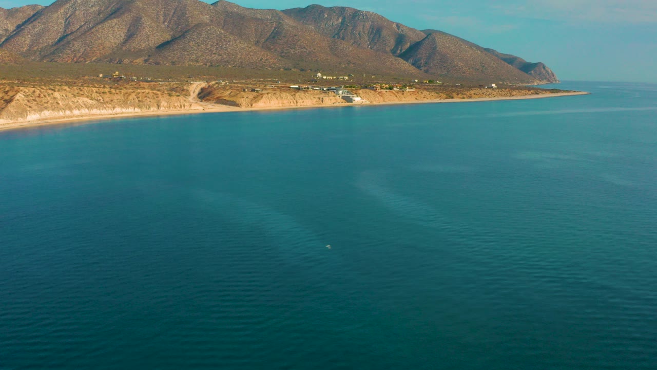 Opening frame over teal water facing the El Sargento La Ventana coastline, with a pale shore, scattered coastal structures, dry mountain slopes, and no visible people.