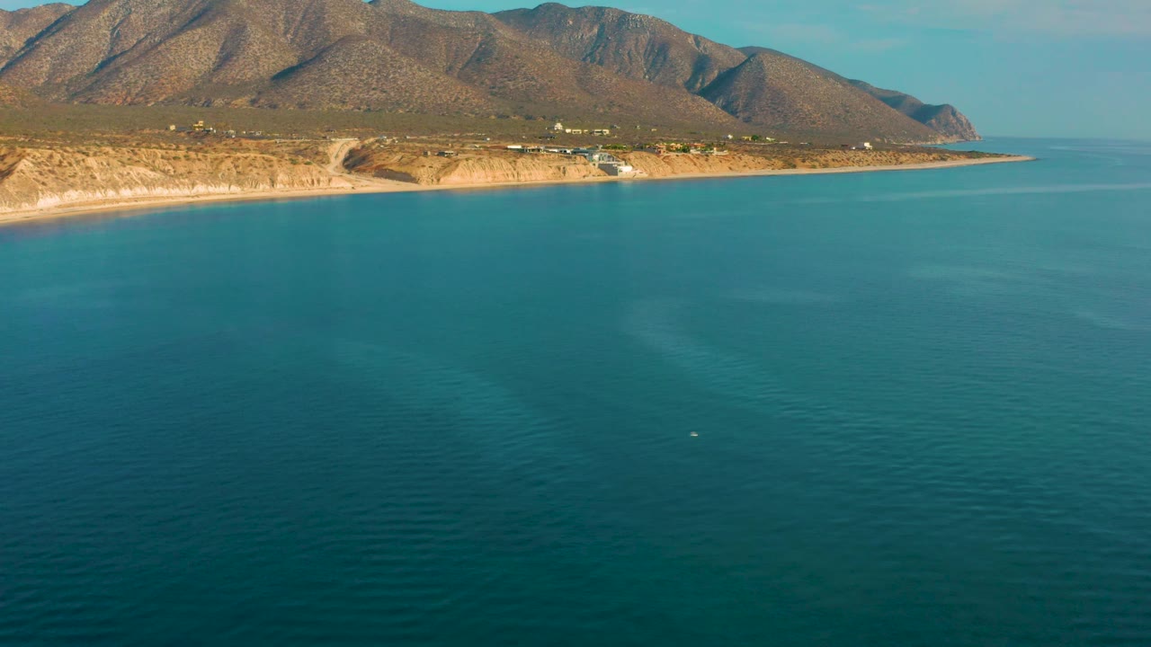 The frame shows turquoise water stretching toward the El Sargento La Ventana shoreline, with tan coastal land, a few small structures, mountain ridges, and no visible people.