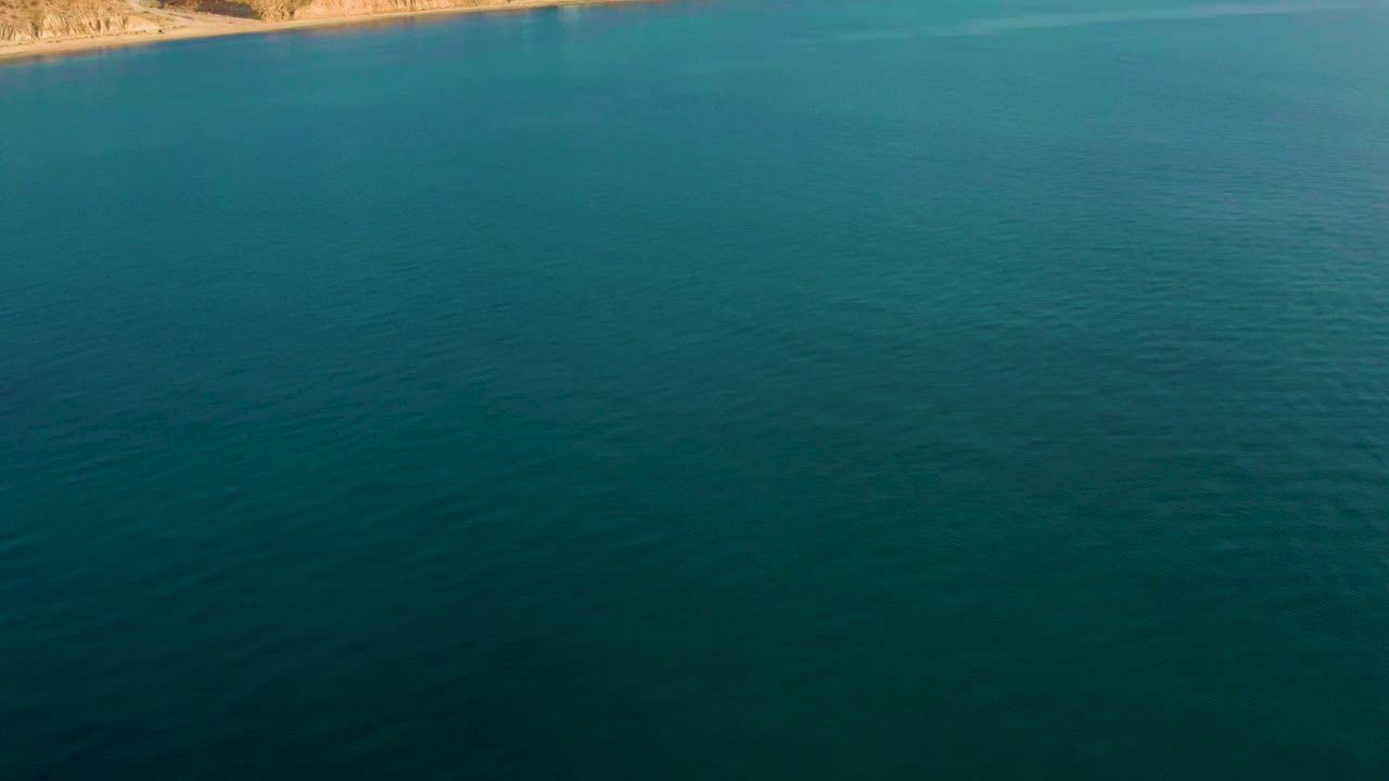 A high aerial view over teal-blue water in El Sargento La Ventana, Mexico, with a thin strip of tan rocky coast along the top edge and no visible people.