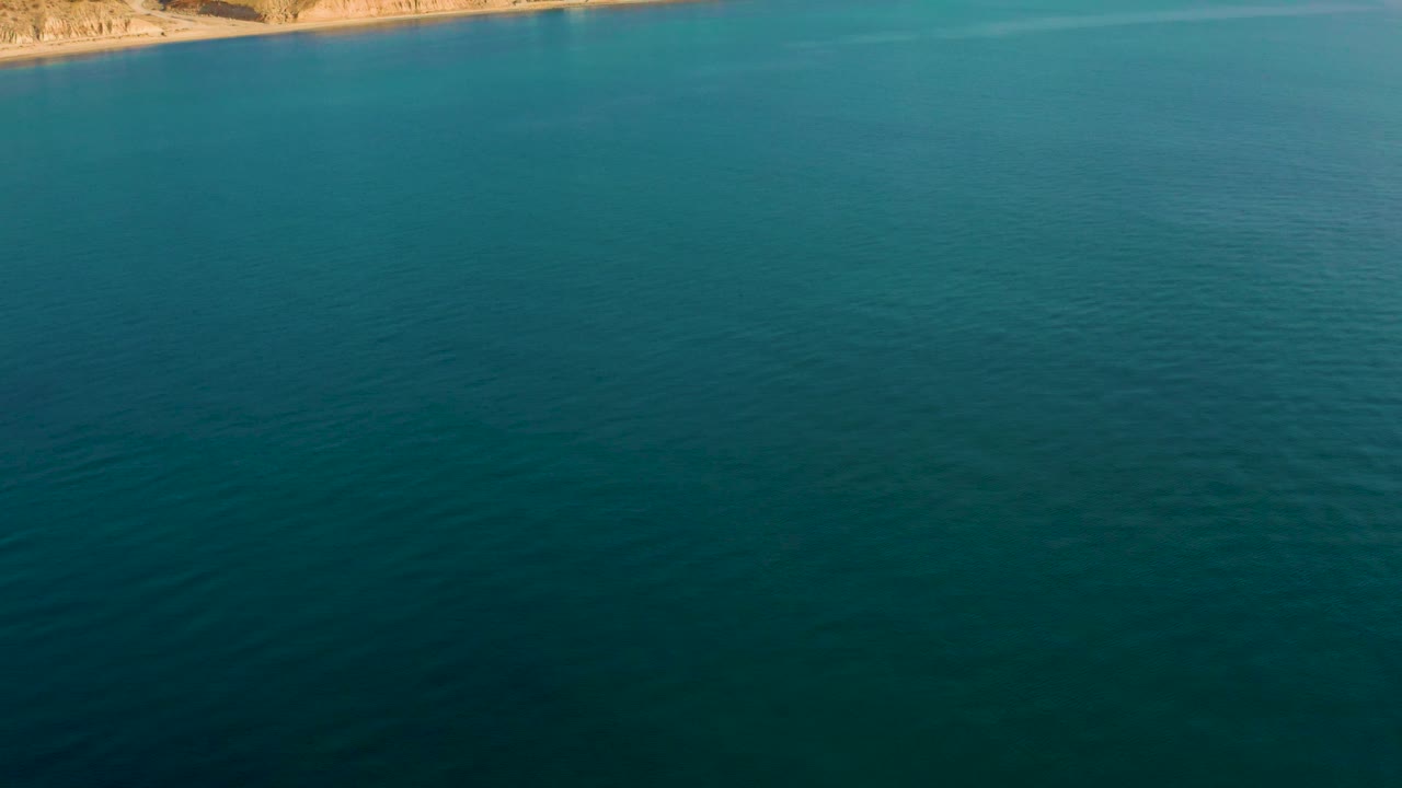 An elevated view over darker teal foreground water in El Sargento La Ventana, Mexico, with faint wave texture and a tan coastal strip at the top, with no visible people.
