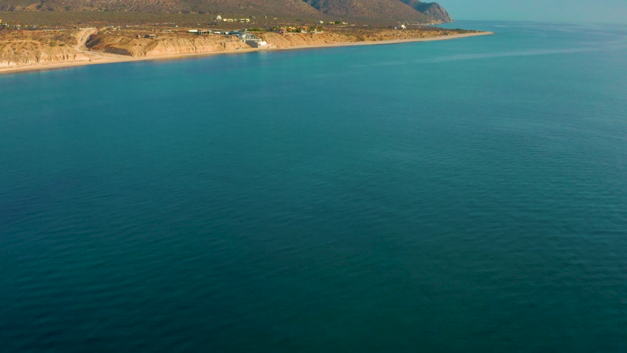 Opening aerial frame over turquoise water facing the El Sargento La Ventana shoreline, with brown mountains behind the coast and no visible people.