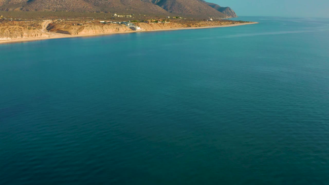 At 0:02, the frame holds a wide drone view of calm ocean leading to the El Sargento La Ventana shoreline and arid mountain ridges, with no visible people.