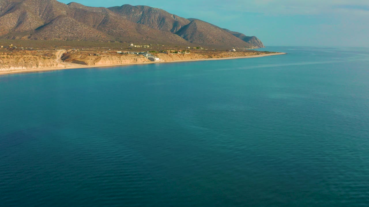A wide aerial frame over El Sargento La Ventana, Mexico shows teal water in front, a thin tan shore with distant buildings, dry mountains behind it, and no visible people.