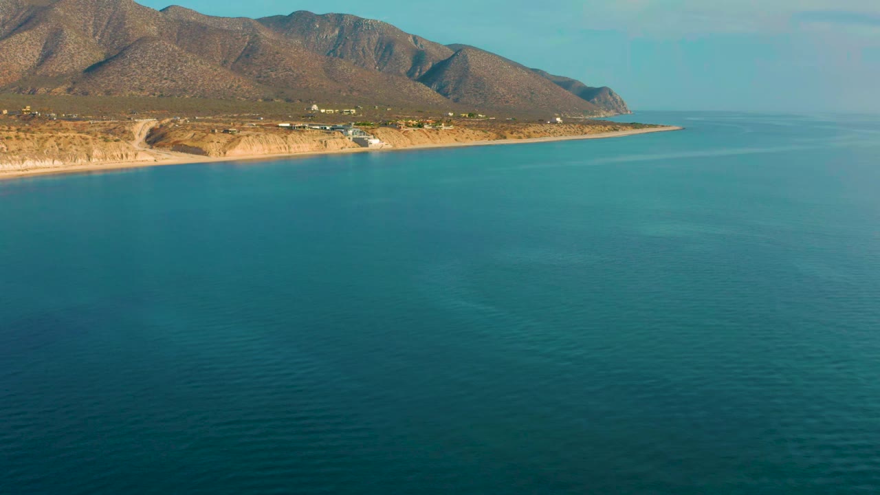 The second frame keeps El Sargento La Ventana coastline high in view, with calm teal water below, scattered shoreline structures, brown mountain ridges, and no visible people.