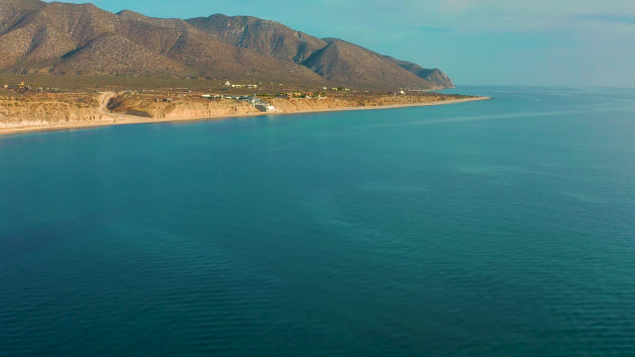 Aerial footage over El Sargento La Ventana shows the coastline stretching left to right, blue-green water occupying most of the frame, rugged hills inland, and no visible people.