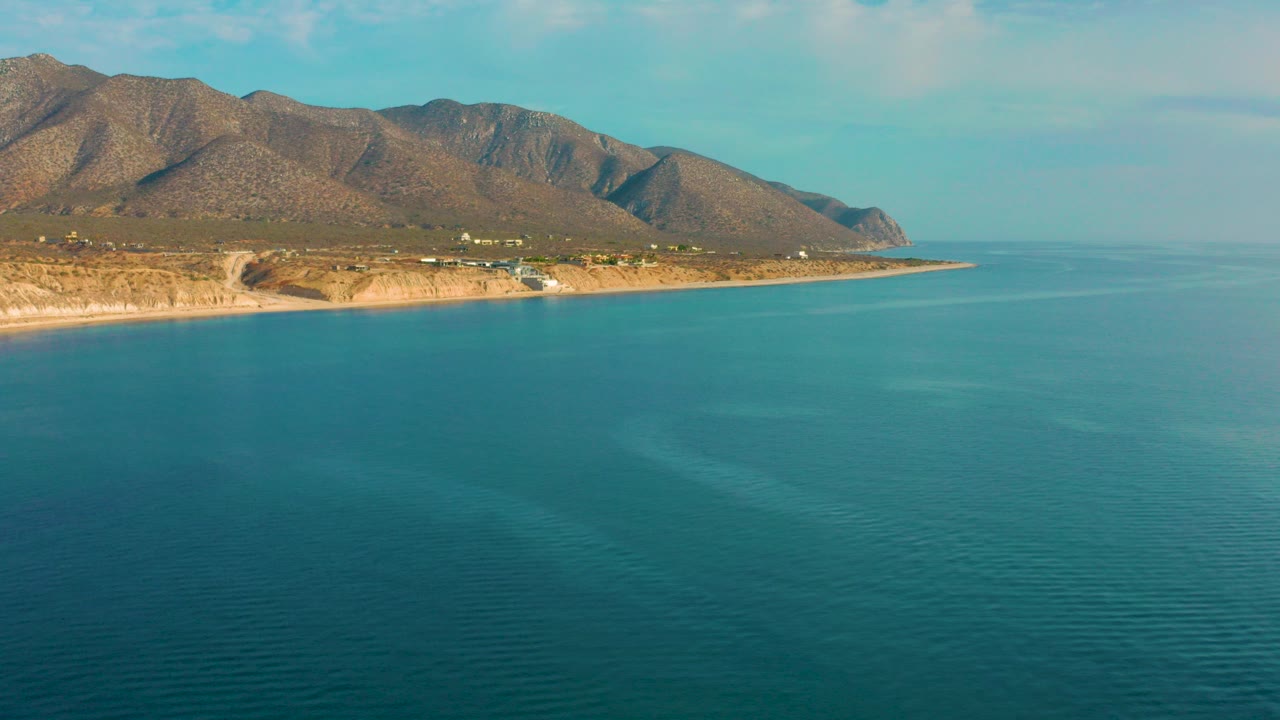 Wide aerial frame over turquoise water facing the El Sargento La Ventana, Mexico shoreline, with tan rocky beach, hillside terrain, distant mountains, and no visible people.