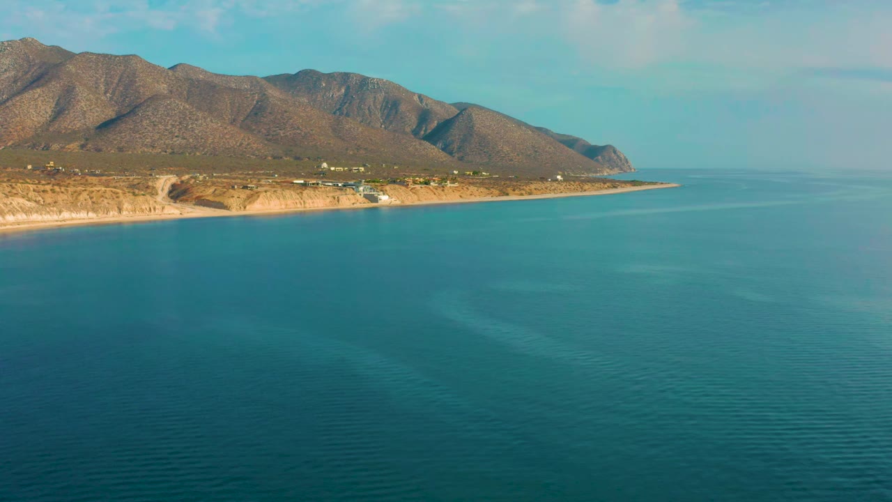 Aerial view in El Sargento La Ventana, Mexico with blue-green sea covering most of the frame, the rocky coast running left to right, mountains behind it, and no visible people.