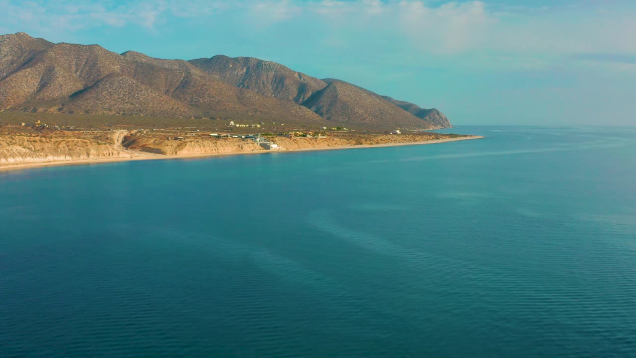 The frame shows the El Sargento La Ventana coast from above, with calm turquoise water, a narrow tan beach, scattered small structures near the shore, mountains beyond, and no visible people.