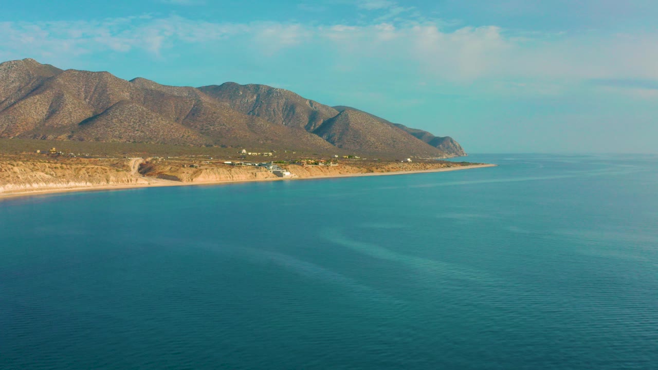 Opening frame of El Sargento La Ventana, Mexico, showing turquoise water in the foreground, a sandy coast with small buildings, brown mountain ridges, pale sky, and no visible people.