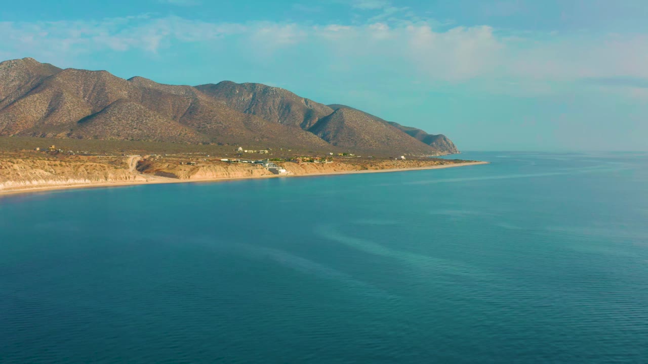 Second frame over El Sargento La Ventana, Mexico, with blue-green ocean filling the lower frame, a thin sandy shoreline, scattered coastal structures, mountains, and no visible people.