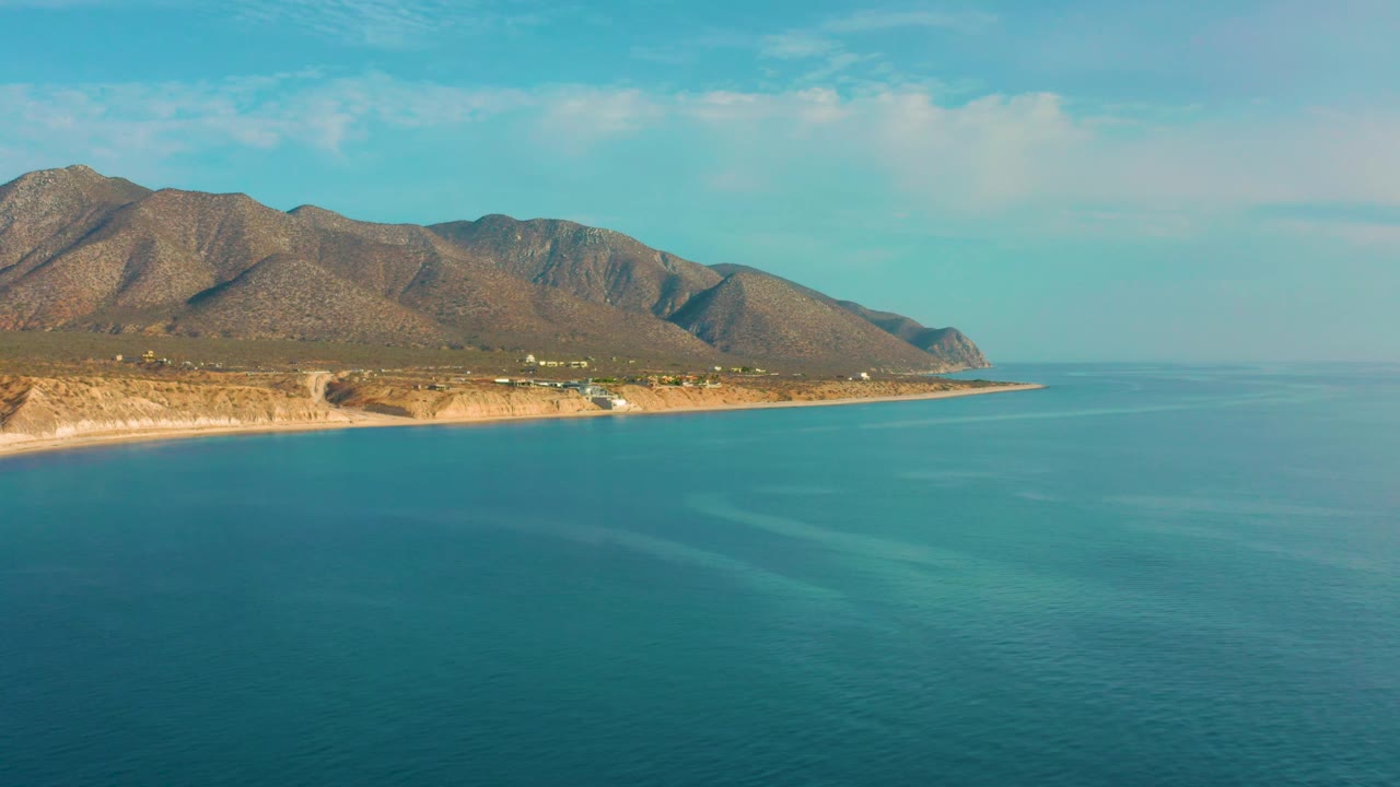 The frame shows faint surface bands across the blue sea, a narrow coastline, and the mountain backdrop at El Sargento La Ventana, Mexico, with no visible people.