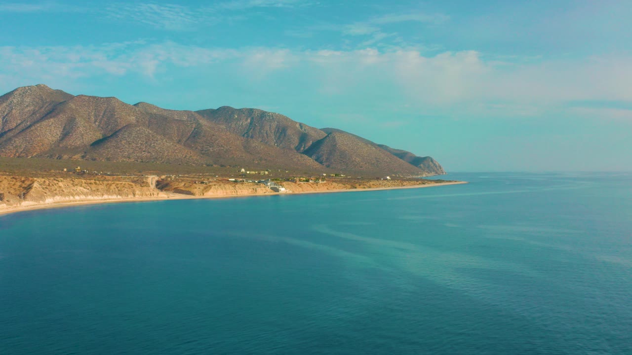 Opening view over El Sargento La Ventana, Mexico with blue sea in the foreground, a tan coastline at left, dry mountains behind, and no visible people.