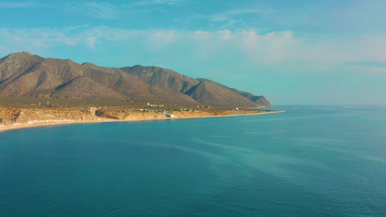 El Sargento La Ventana coastline remains high in frame, with turquoise water below, a mountain ridge across the background, and no visible people.