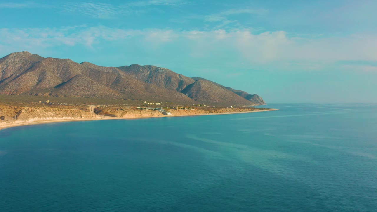 A broad blue ocean view faces the tan coast of El Sargento La Ventana, Mexico, with rocky mountains under a pale sky and no visible people.